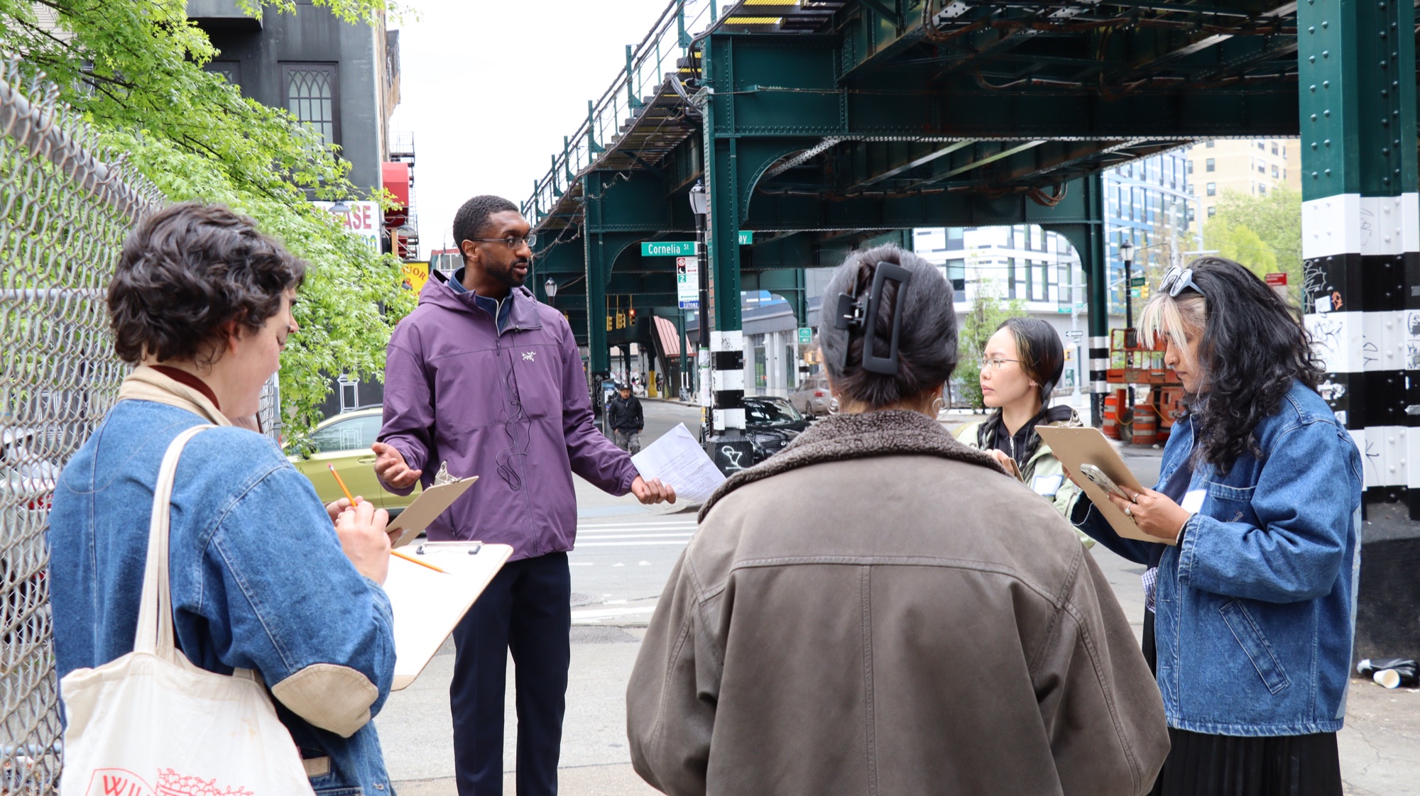 The guide talks through the problem with walkers holding clipboards under the elevated train at Broadway and Cornelia St