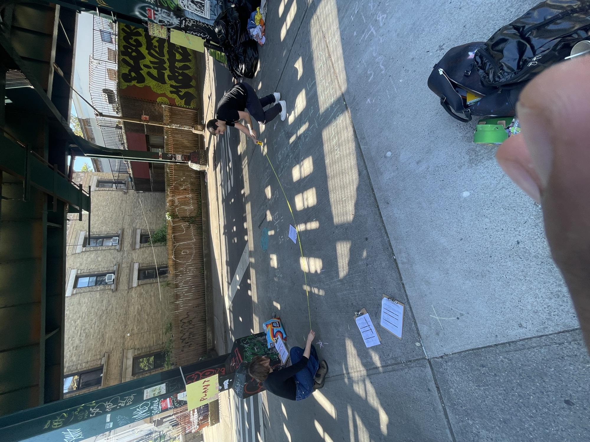 Two walkers stretching a tape measure across the pavement under the elevated M, chalk math on the ground